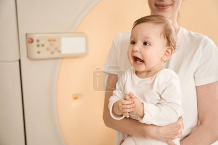 Foto de Foto recortada de madre alegre con niño pequeño en brazos de pie delante de la máquina de resonancia magnética en la sala de exploración - Imagen libre de derechos
