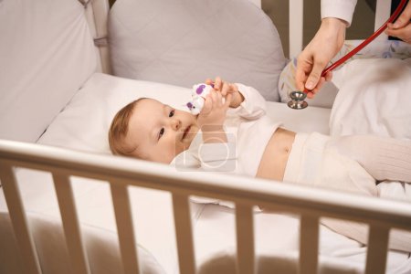 Téléchargez les photos : Pédiatre avec stéthoscope atteignant pour bébé nouveau-né calme couché dans un lit avec jouet dans les mains - en image libre de droit