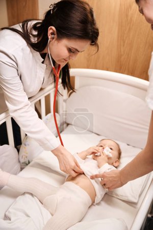 Téléchargez les photos : Pédiatre féminine ciblée à l'écoute des battements de coeur du nouveau-né avec stéthoscope en présence du parent - en image libre de droit