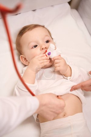 Téléchargez les photos : Calme bébé mettre jouet dans la bouche pendant l'auscultation cardiaque effectuée par un pédiatre avec l'aide des parents - en image libre de droit