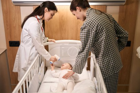 Téléchargez les photos : Médecin pédiatrique ciblé pointant du doigt un thermomètre infrarouge sans contact sur le front du nouveau-né en présence d'une mère heureuse - en image libre de droit