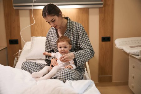 Foto de Mujer joven en pijama sentada con un niño enfermo en la cama en la sala de hospital - Imagen libre de derechos