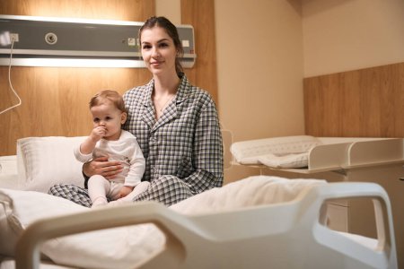 Joven mamá en pijama sentada con su bebé enfermo en la cama en la sala de hospital