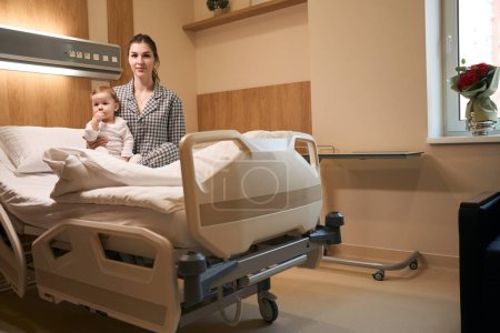 Téléchargez les photos : Femme calme vêtue de pyjama assise avec un enfant malade sur le lit dans la salle - en image libre de droit