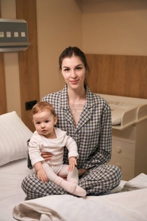 Téléchargez les photos : Femme souriante en pyjama assise avec bébé malade sur le lit dans la salle - en image libre de droit