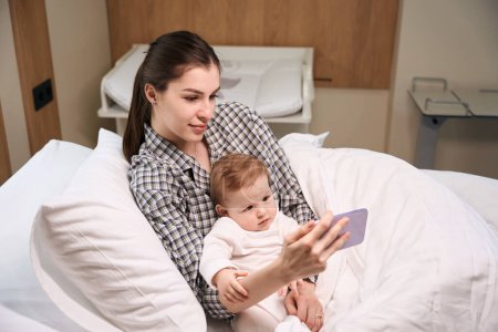 Foto de Madre mujer mostrando dibujos animados en el teléfono móvil a su bebé acostado en la cama en la sala - Imagen libre de derechos