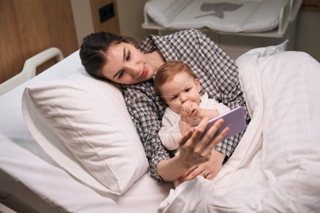 Foto de Mujer joven y niño viendo vídeo en el teléfono inteligente mientras está acostado en la cama del hospital - Imagen libre de derechos