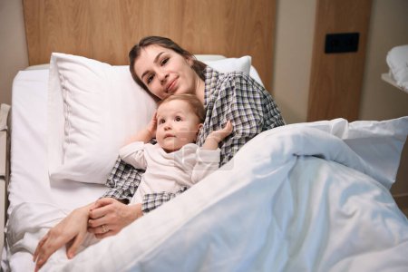 Téléchargez les photos : Jeune femme souriante couchée avec son petit bébé malade dans un lit d'hôpital - en image libre de droit