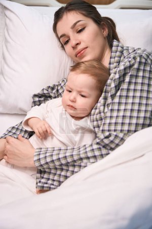 Foto de Pensativo padre femenino acostado con su triste hijo enfermo en la cama - Imagen libre de derechos