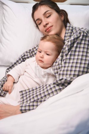Foto de Mujer joven y pacífica acostada con un bebé enfermo en la cama envuelto en edredón - Imagen libre de derechos
