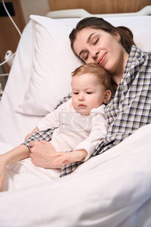Foto de Mujer sonriente acostada junto a su triste hijita en la cama del hospital - Imagen libre de derechos