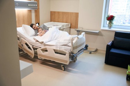 Téléchargez les photos : Femme lisant les nouvelles sur le téléphone portable tout en couchant avec son bébé malade dans le lit d'hôpital - en image libre de droit