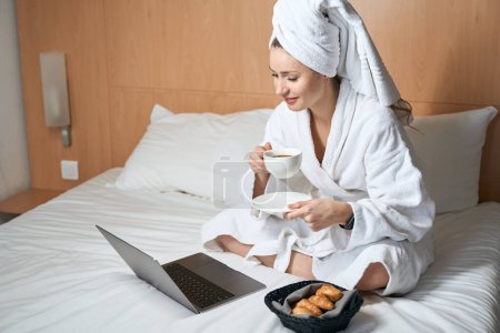 Foto de Foto de mujer en albornoz con laptop bebiendo café con croissants en la cama en la habitación del hotel - Imagen libre de derechos