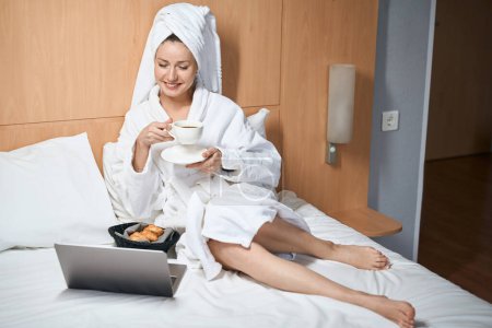 Foto de Retrato de una hermosa dama con un portátil bebiendo café con croissants sentado en la cama en la habitación del hotel - Imagen libre de derechos