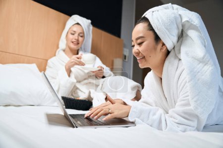 Téléchargez les photos : Photo de deux amies se reposant sur le lit en peignoirs blancs sur le lit de l'hôtel - en image libre de droit