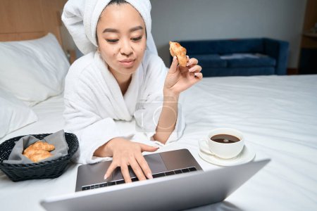 Foto de Mujer asiática en bata blanca usando portátil en la cama. Lady come croissants con café en la cama del hotel - Imagen libre de derechos
