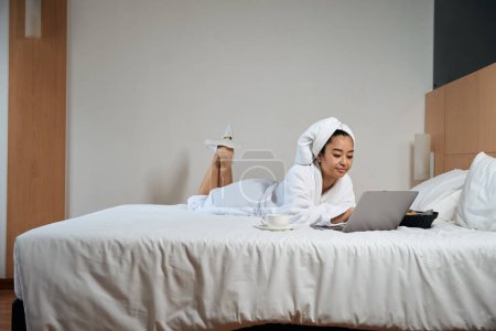 Téléchargez les photos : Photo de femme asiatique sur le lit en peignoir avec ordinateur portable. Dame en peignoir avec ordinateur portable sur le lit dans la chambre d'hôtel - en image libre de droit