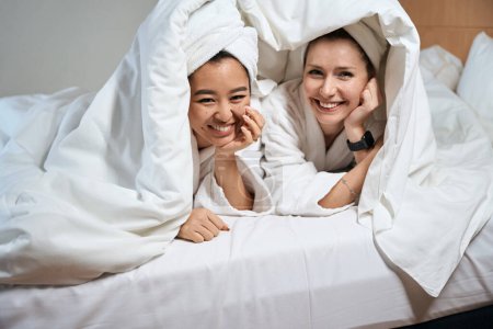 Téléchargez les photos : Mignonnes dames jettent un coup d'oeil sous les couvertures sur le lit. Ami joyeux se cachent sous la couverture sur le lit - en image libre de droit