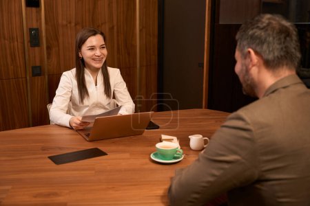 Téléchargez les photos : Jeune nutritionniste féminine assise devant un homme adulte au bureau avec un ordinateur portable et tenant la prescription dans ses mains - en image libre de droit