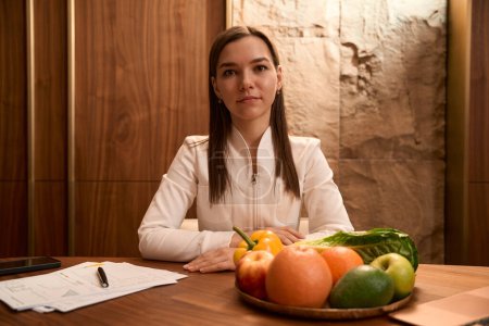Téléchargez les photos : Jeune nutritionniste assis les mains croisées au bureau avec prescription et légumes au bureau - en image libre de droit