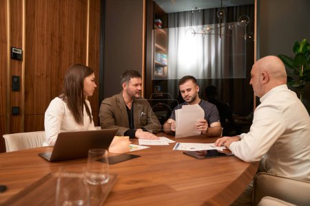 Photo for Young physician sitting at desk with colleagues and presenting papers to man in the office - Royalty Free Image