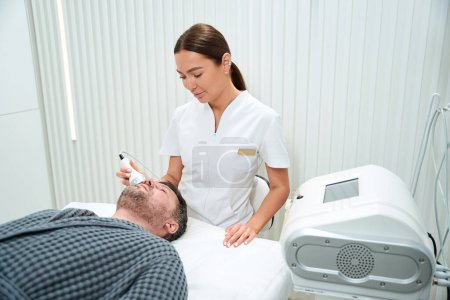 Téléchargez les photos : Jeune cosmétologue debout à côté d'un patient couché et faisant une intervention sur le visage dans le salon de beauté - en image libre de droit