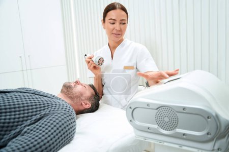 Téléchargez les photos : Infirmière debout près de l'homme couché et dispositif de maintien pour soulever RF dans sa main dans le spa - en image libre de droit