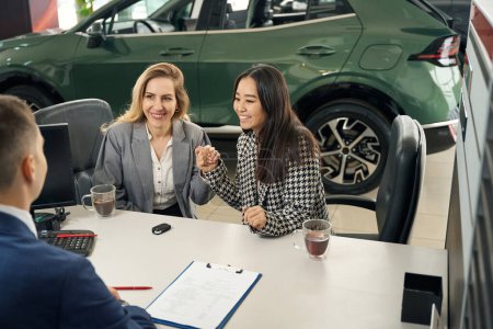 Foto de Feliz pareja LGBT femenina está solicitando un préstamo de coche en un concesionario de automóviles, un gerente está hablando con ellos - Imagen libre de derechos