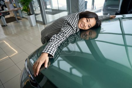 Glückliche junge asiatische Frau umarmt die Motorhaube eines neuen Autos, sie trägt bequeme Freizeitkleidung