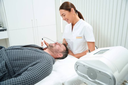 Foto de Bastante cosmetólogo realiza un procedimiento de elevación de RF en un hombre de mediana edad, el especialista utiliza un dispositivo moderno - Imagen libre de derechos