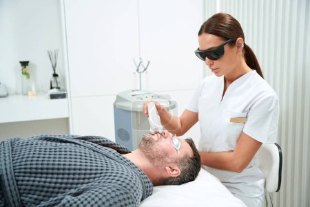 Foto de Mujer con gafas realiza un procedimiento de fotorejuvenecimiento en un hombre de mediana edad, la paciente se encuentra en un sofá de cosmetología - Imagen libre de derechos