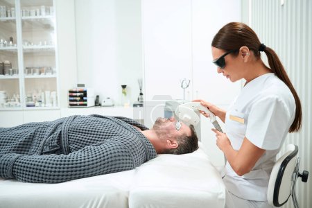 Foto de Paciente se encuentra en un sofá de cosmetología durante el procedimiento de fotorejuvenecimiento, el cosmetólogo trabaja con el área frontal - Imagen libre de derechos