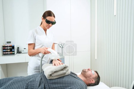Foto de Cosmetólogo realiza un procedimiento de fotorejuvenecimiento en las manos de los pacientes, el hombre se encuentra en un sofá de cosmetología - Imagen libre de derechos