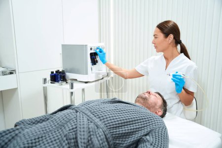 Téléchargez les photos : Cosmetologue ajuste le dispositif pour la procédure de microdermabrasion laser rajeunissante, le patient se trouve sur le canapé - en image libre de droit