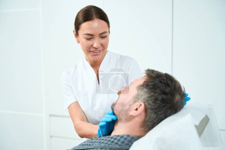 Téléchargez les photos : Esthéticienne mignonne examine le visage d'un patient d'âge moyen, un homme confortablement installé sur un canapé de cosmétologie - en image libre de droit