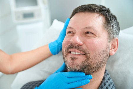 Téléchargez les photos : Travailleur de la santé dans des gants de protection examine le visage d'un patient d'âge moyen, un homme est situé sur un canapé de cosmétologie - en image libre de droit