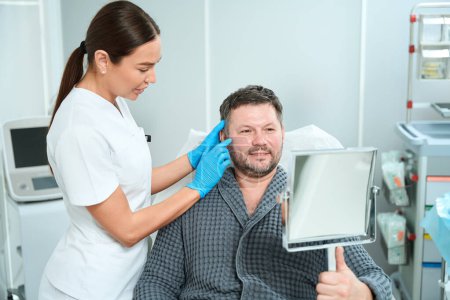 Téléchargez les photos : Homme souriant assis devant un miroir lors d'une consultation avec un esthéticien, un spécialiste examine le visage des patients - en image libre de droit