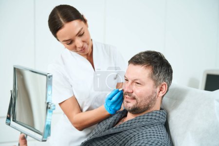 Photo for Man sits in front of a mirror at a consultation with a cosmetologist, a specialist applies markers to patients face - Royalty Free Image