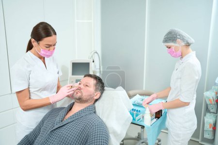 Photo for Middle-aged patient on a rejuvenating plasma therapy procedure, male receives subcutaneous injections, doctor and assistant in protective masks - Royalty Free Image