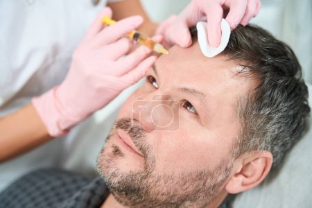 Foto de Hombre con la cara sin afeitar recibe inyecciones subcutáneas de la zona frontal, un médico en guantes de protección - Imagen libre de derechos