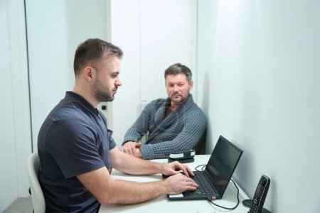Foto de Nutricionista masculino está escribiendo en un ordenador portátil en el lugar de trabajo, que está consultando a un paciente masculino en una bata de baño - Imagen libre de derechos