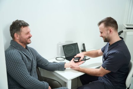 Téléchargez les photos : Homme barbu dans un centre médical subit une procédure de diagnostic oligoscan, un spécialiste utilise un appareil spécial - en image libre de droit
