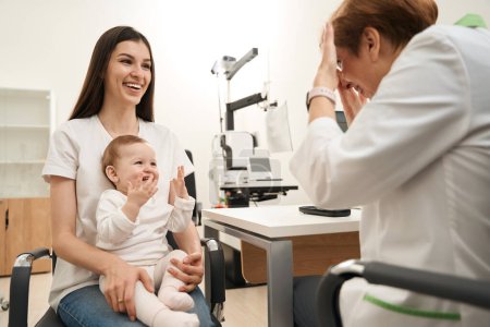Téléchargez les photos : Ophtalmologiste pédiatrique attirant l'attention du bébé souriant avec un objet intéressant en présence de la mère heureuse lors de l'examen de la vue - en image libre de droit