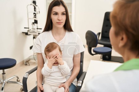 Photo for Serious woman seated with child in chair listening to pediatric eye doctor - Royalty Free Image
