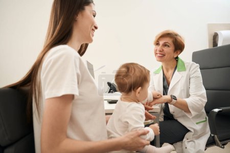 Téléchargez les photos : Joyeuse ophtalmologiste pédiatrique communiquant avec une jeune maman heureuse assise sur une chaise avec un bébé dans les bras - en image libre de droit
