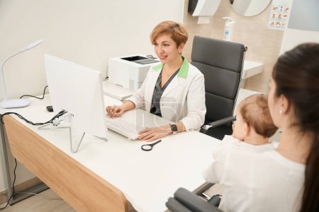 Téléchargez les photos : Médecin généraliste souriant assis au bureau saisissant des informations dans l'ordinateur en présence de la mère et de l'enfant lors de la consultation initiale - en image libre de droit