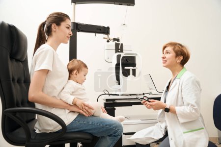 Photo for Smiling pediatric ophthalmologist with magnifiers in hands looking at woman with baby seated in her office - Royalty Free Image
