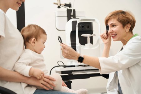 Téléchargez les photos : Ophtalmologiste pédiatrique agréable vérifiant la vision du nourrisson avec occlusion et loupe lors de la consultation - en image libre de droit