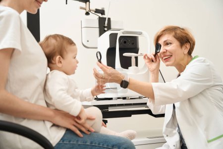 Photo for Joyous pediatric optometrist checking baby eyesight with magnifier and occluder in mother presence - Royalty Free Image