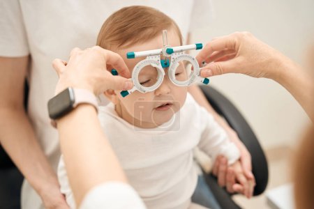 Téléchargez les photos : Des mains de ophtalmologistes portant un cadre d'essai ophtalmique sur un petit enfant calme en présence de parents - en image libre de droit